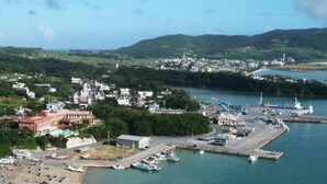 A coastal town with a marina, buildings, and a hilly landscape in the background. - Hotel Garden Hills (Kumejima)