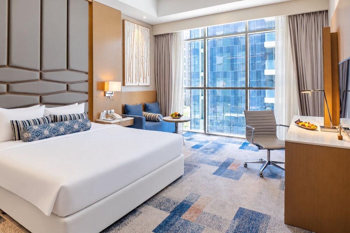 A hotel room with a large bed, a desk, a chair, a small table, and a view of the city through large windows.