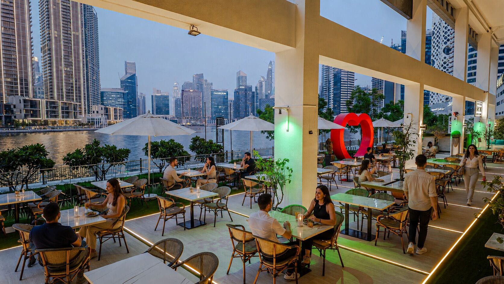 An outdoor dining area with tables and chairs, a view of the city skyline, and a large red sculpture.