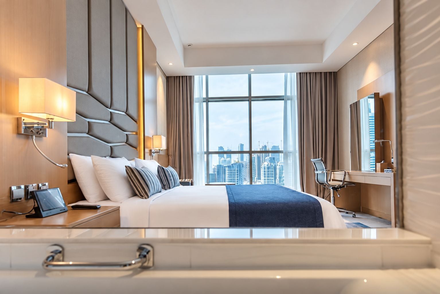 A modern hotel room with a large bed, city view, and a desk with a chair.