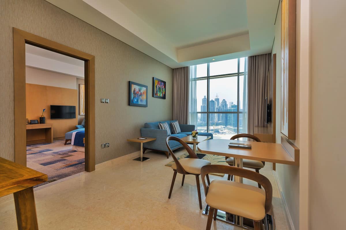 A modern hotel room with a city view, a sofa, a dining table with chairs, and a TV.