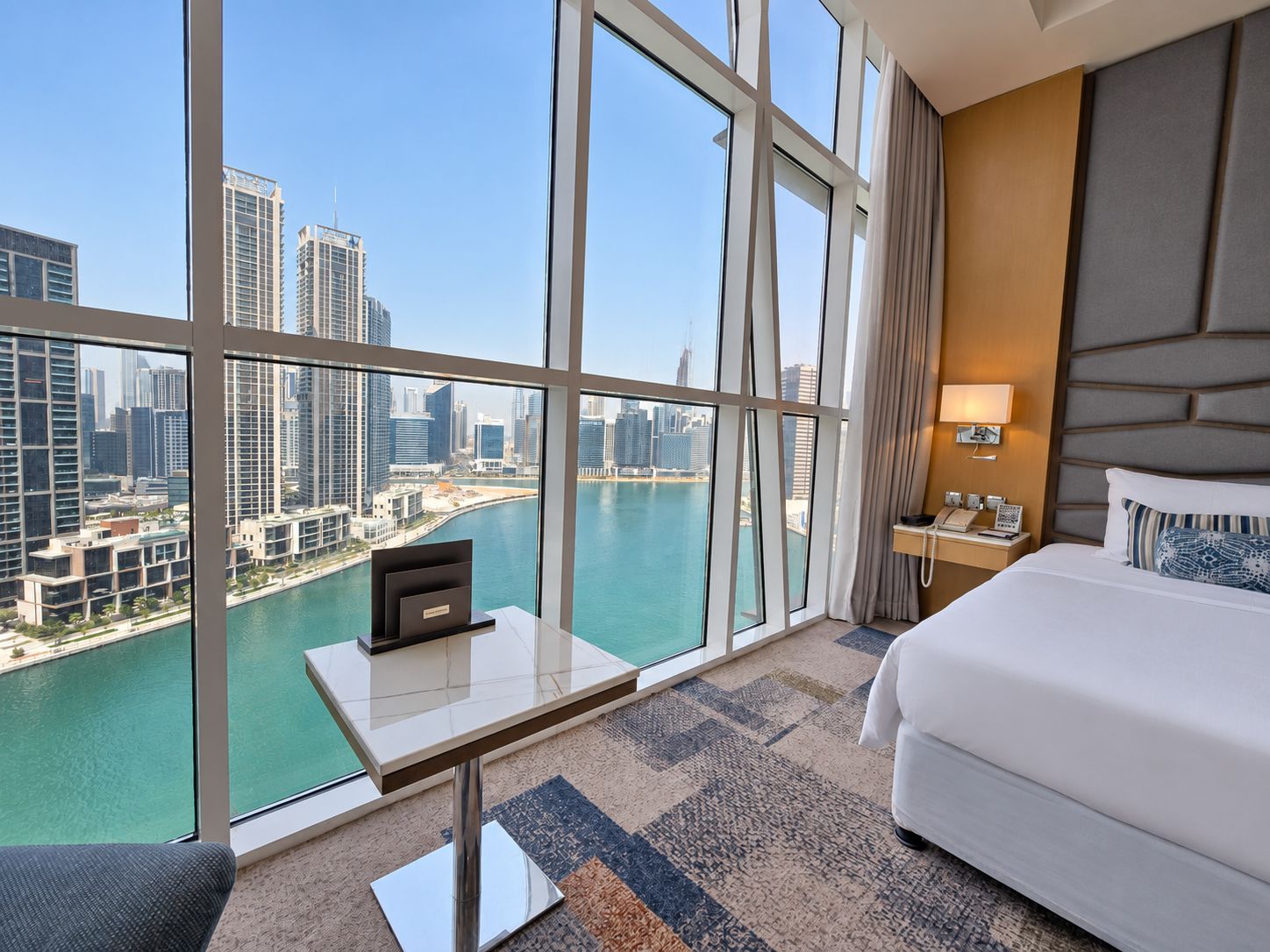 A hotel room with a large window, a bed, a bedside table with a lamp, and a view of a cityscape and water.