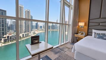 A hotel room with a large window, a bed, a bedside table with a lamp, and a view of a cityscape and water.