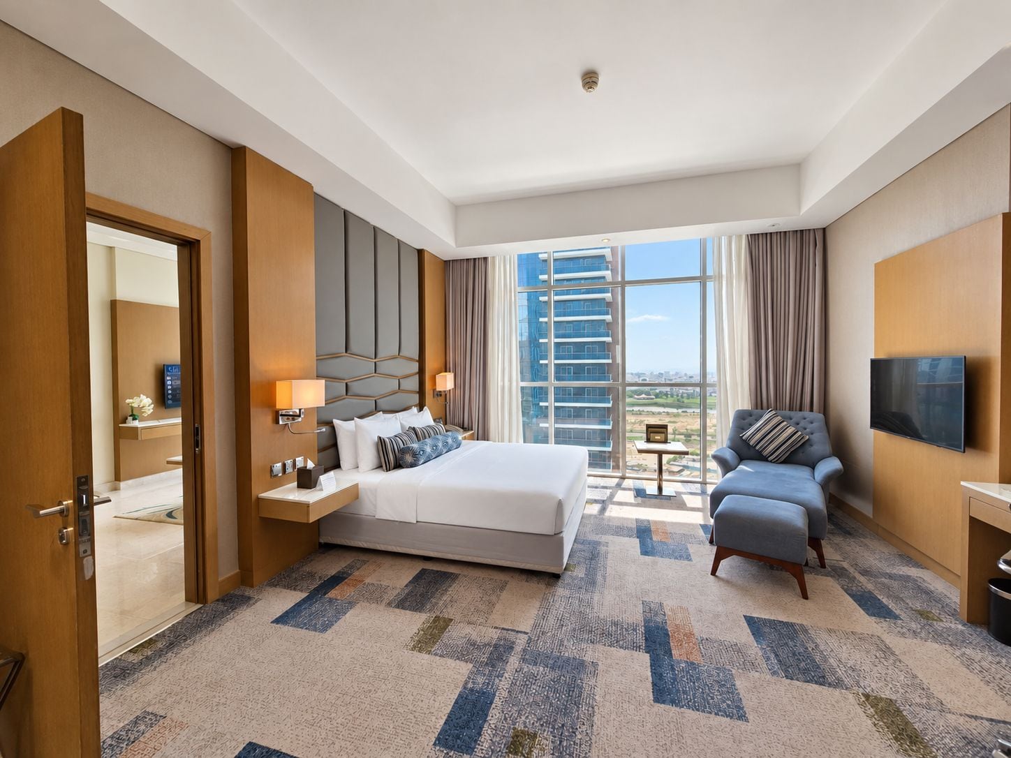 A hotel room with a large bed, a sofa, a TV, and a view of a high-rise building.