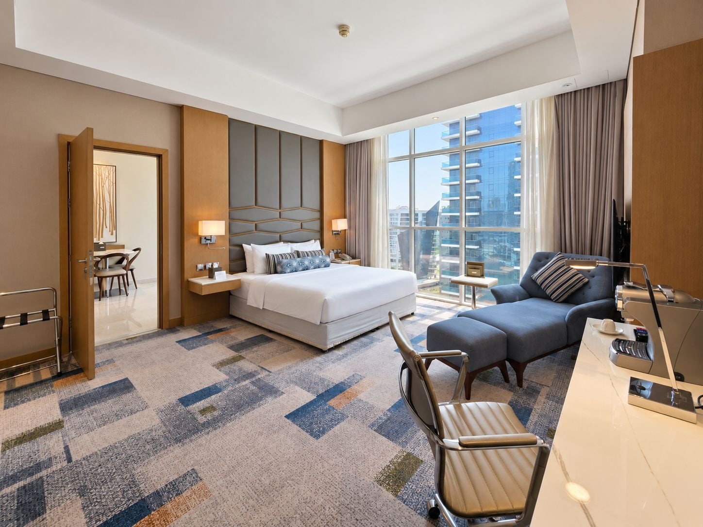 A hotel room with a large bed, a sofa, a desk, and a view of a cityscape through large windows.