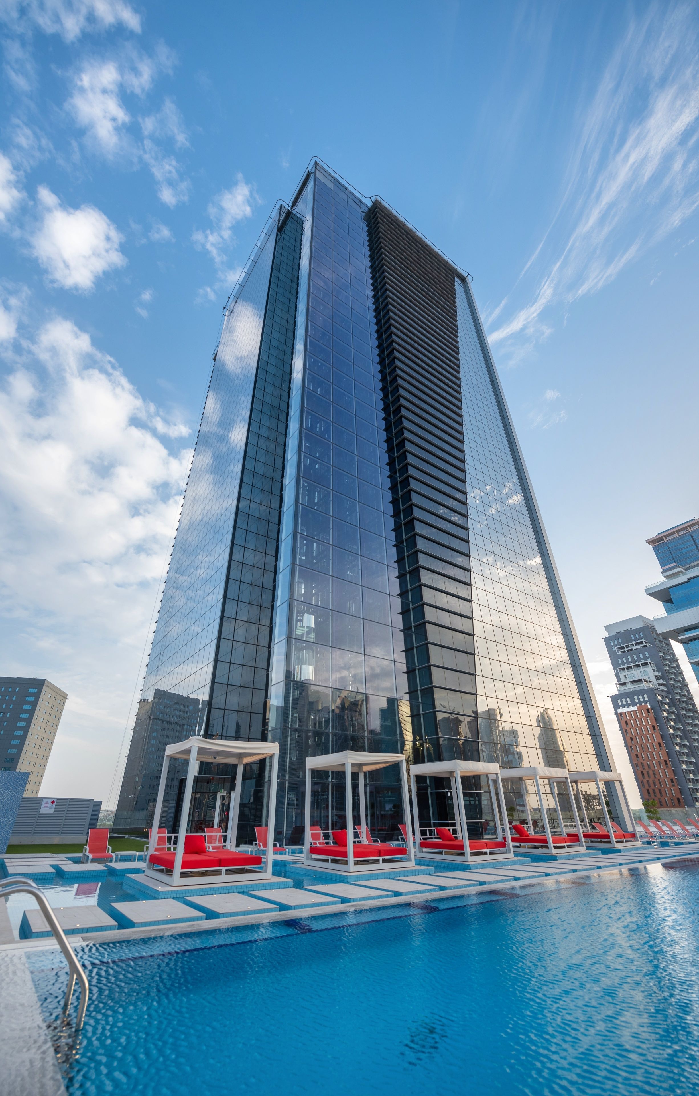A modern high-rise building with a reflective glass facade, a rooftop pool with red lounge chairs, and a clear blue sky.