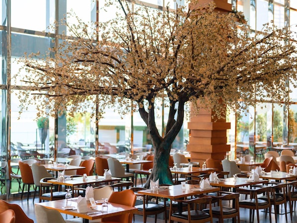 A restaurant with tables set for dining, a large tree with blossoms in the center, and large windows in the background. - Canal Central Hotel (Dubai)