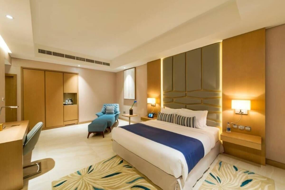 A hotel room with a bed, a desk, a chair, a nightstand, and a wall-mounted lamp.