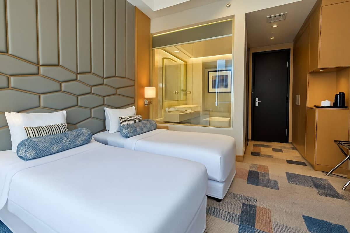 A hotel room with two beds, a glass-enclosed bathroom, and a small seating area.