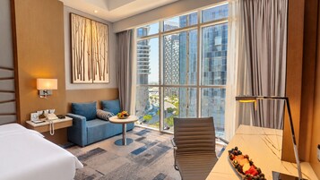 Habitación de hotel con cama, sofá, una pequeña mesa redonda con fruta, escritorio con teléfono y una silla.