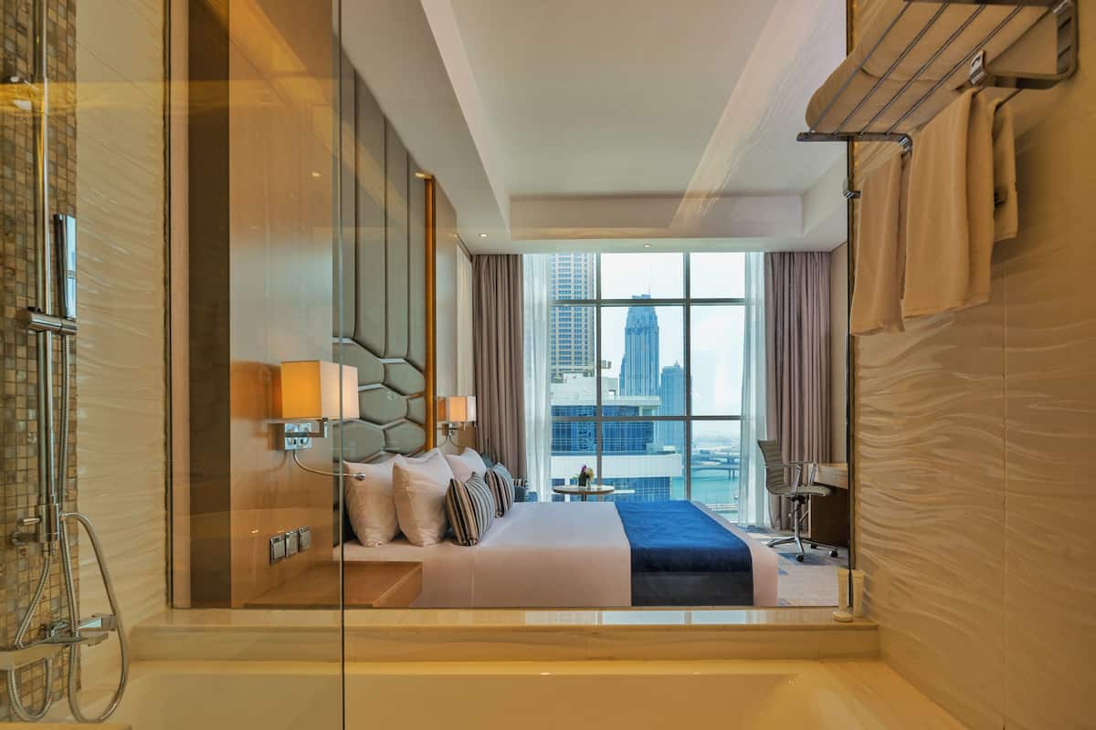 A modern hotel room with a large bed, a glass shower enclosure, and a city view.