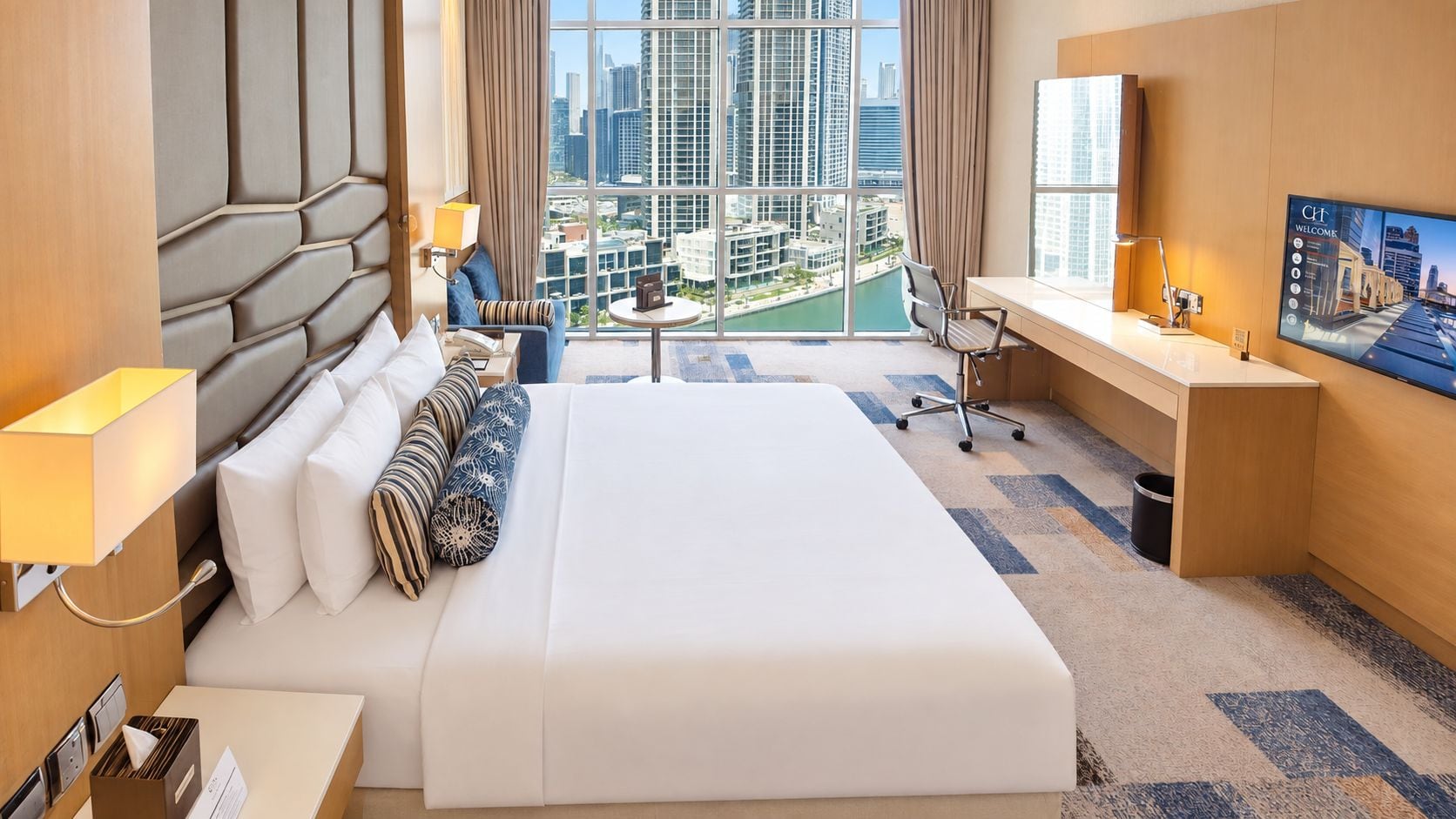 Ein Hotelzimmer mit einem großen Bett, einem Schreibtisch, einem Sessel, einem Fernseher und Blick auf eine Stadtlandschaft.