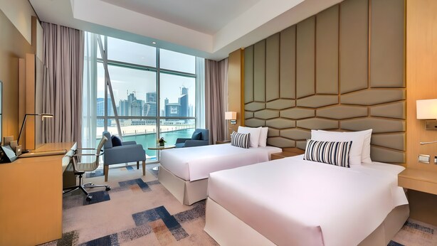 A hotel room with two beds, a desk, a chair, a small table, and a view of the city skyline. - Canal Central Hotel (Dubai)