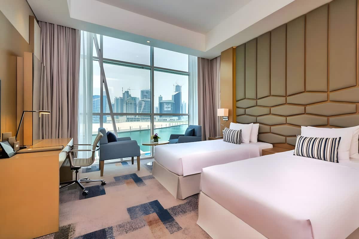 A hotel room with two beds, a desk, a chair, a small table, and a view of the city skyline.