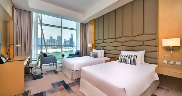 A hotel room with two beds, a desk, a chair, a small table, and a view of the city skyline. - Canal Central Hotel (Dubai)