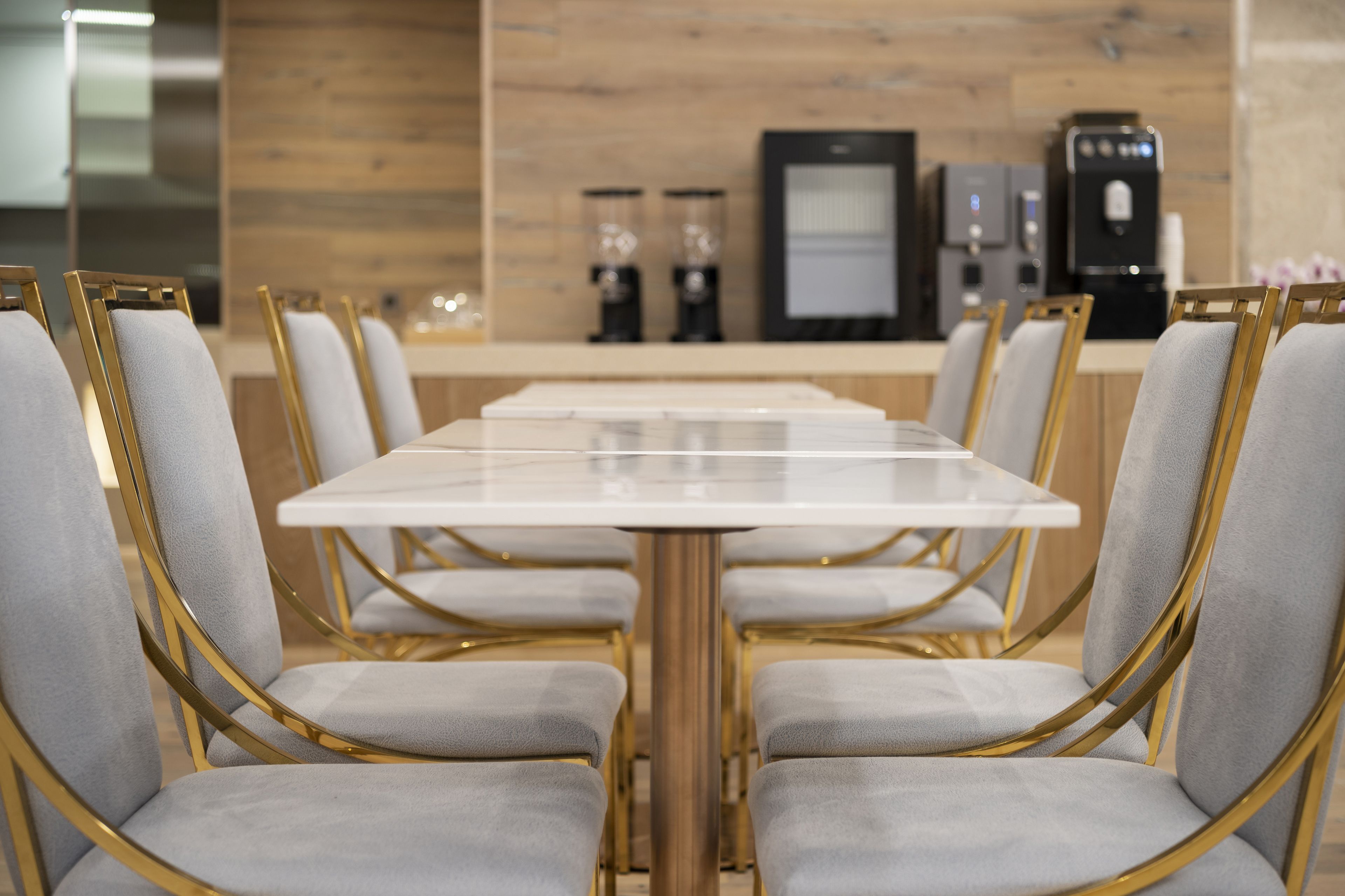 A modern dining area with a long table and chairs, a coffee machine, and a refrigerator in the background.