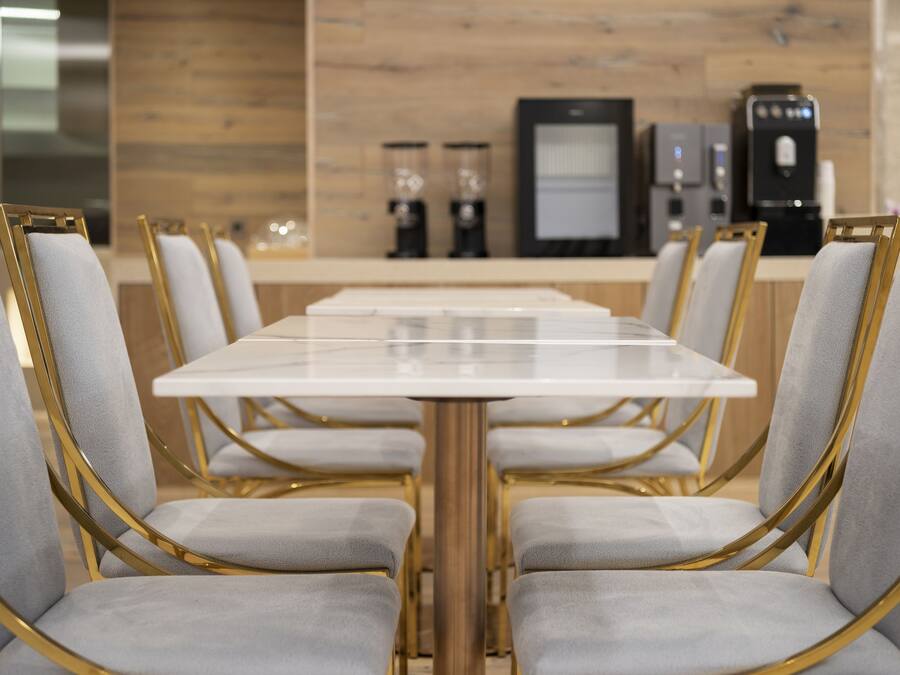 A modern dining area with a long table and chairs, a coffee machine, and a refrigerator in the background.
