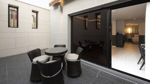 A modern outdoor seating area with wicker chairs and a round table, adjacent to a glass door leading to an interior space with a sofa and a lamp. - Yeonsan Hound Hotel (Busan)