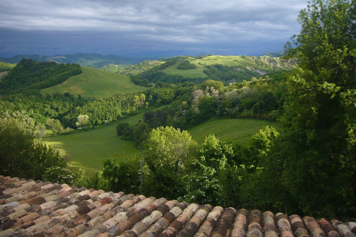 On aperçoit des collines douces et une végétation luxuriante, avec un toit de tuiles au premier plan.