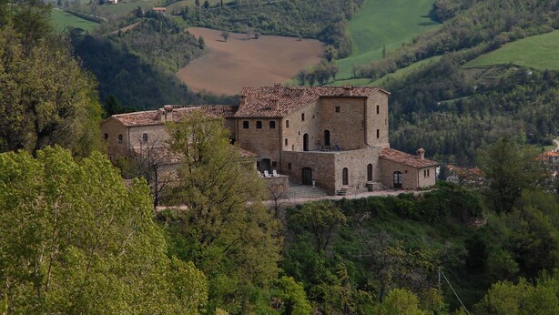 Un edifici de pedra històric amb diverses torres, envoltat per turons verds i un paisatge de vall.