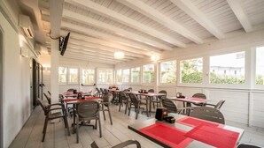 Un restaurante con techo alto y amplios ventanales, que ofrece luz natural y vistas al exterior. Las mesas están dispuestas con individuales rojos, y hay sillas y mesas para comer.
