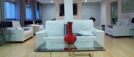 A modern living room with a white sofa, a glass coffee table, and a red vase. There are two white armchairs, a lamp, and a TV on a stand. The room has wooden flooring and patterned wallpaper.