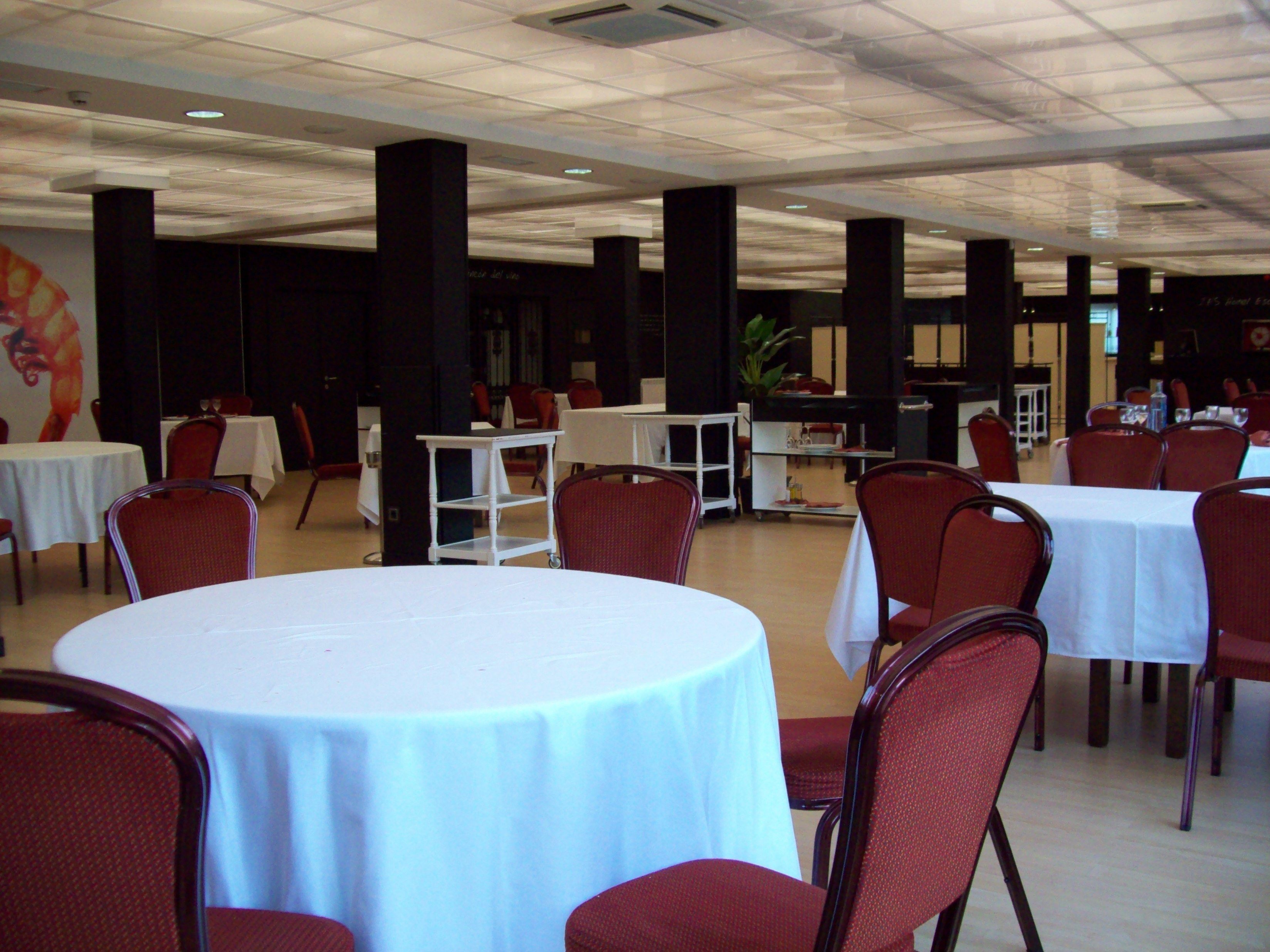 A spacious indoor dining area with round tables and red chairs, featuring a high ceiling with a grid pattern and a large mural on the wall.