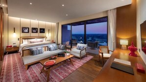 Une chambre d’hôtel moderne, dotée d’un grand lit, d’un canapé, d’une table à manger et offrant une vue sur le paysage urbain au crépuscule.