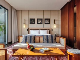 Una habitación de hotel moderna con una cama grande, un sofá, una mesita de centro con un libro y un frutero, y vistas al exterior.