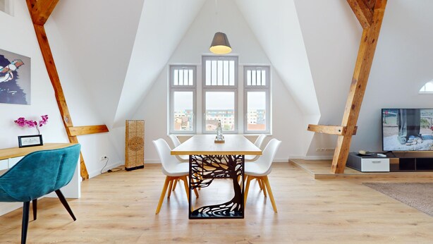 A dining area with a white table and chairs, a decorative tree trunk divider, and a view of buildings through large windows. - Apartment Senftenberg - 1907 (Senftenberg)