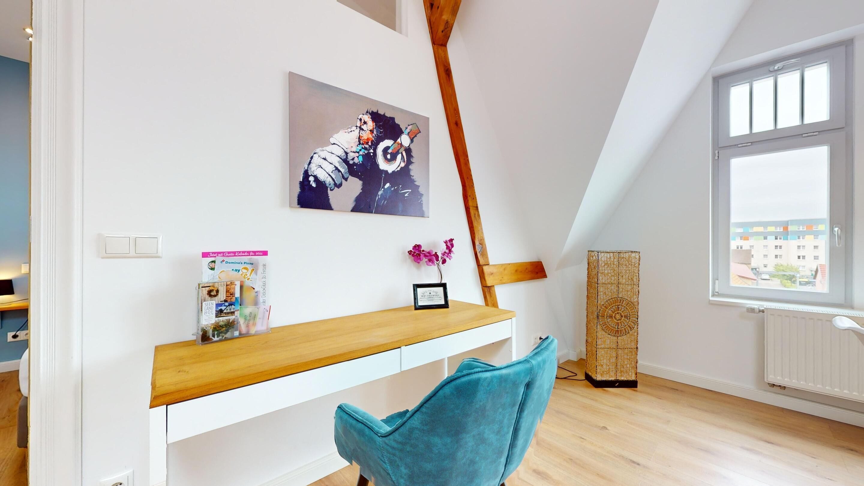 A modern room with a wooden desk, a teal chair, a painting, a framed certificate, and a window with a view of buildings.