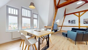 A modern living room with a wooden dining table, white chairs, a blue sofa, and a TV. Large windows offer a view of buildings outside. - Apartment Senftenberg - 1907 (Senftenberg)
