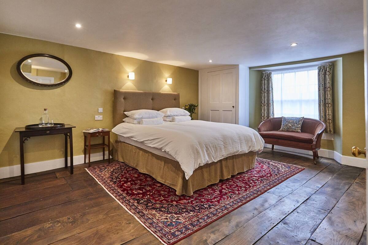 A bedroom with a large bed, a nightstand, a chair, a mirror, and a window with curtains.
