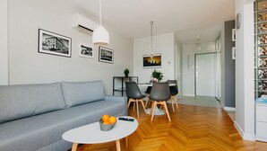 Een moderne woonkamer met een grijze bank, een ronde salontafel met mandarijnen, een eethoek met een kroonluchter, en ingelijste foto’s aan de muur.