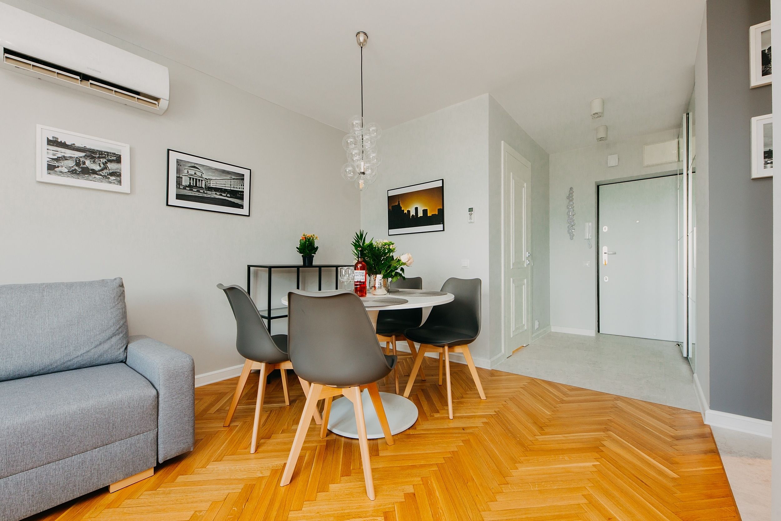 Una sala de estar moderna con un sofá gris, una mesa de comedor con sillas, una araña y un piso de madera con patrón de espiga.