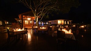 An outdoor dining area with tables and chairs, illuminated by candlelight, under a large tree.