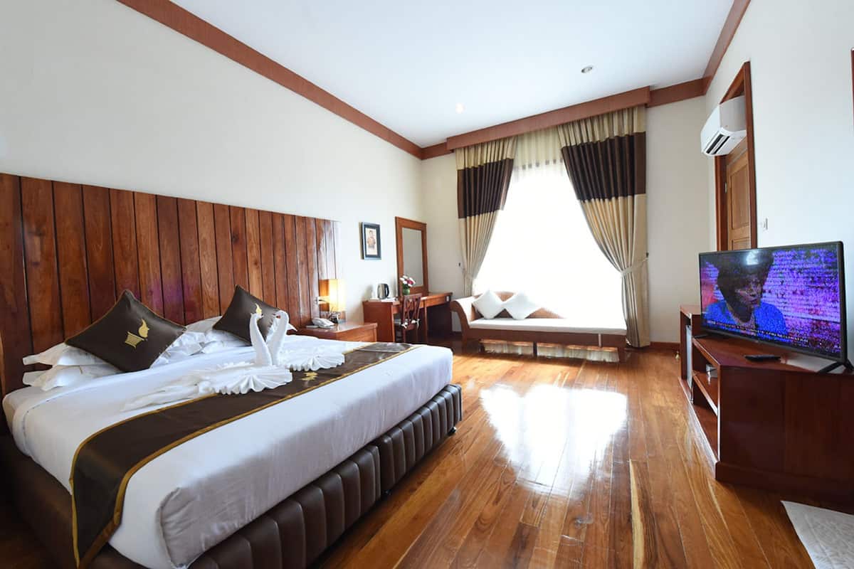 A hotel room with a large bed, wooden flooring, a television, and a window with curtains.