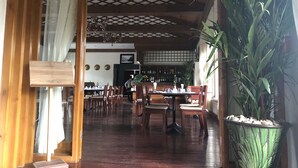 A restaurant with wooden flooring, tables set with white tablecloths, and chairs. There is a large potted plant on the right.
