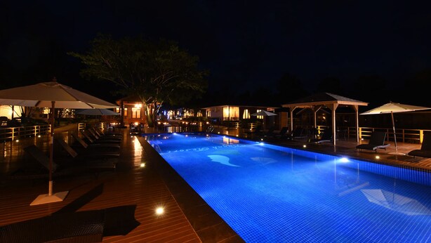 A nighttime swimming pool with lounge chairs and umbrellas, surrounded by buildings and trees.