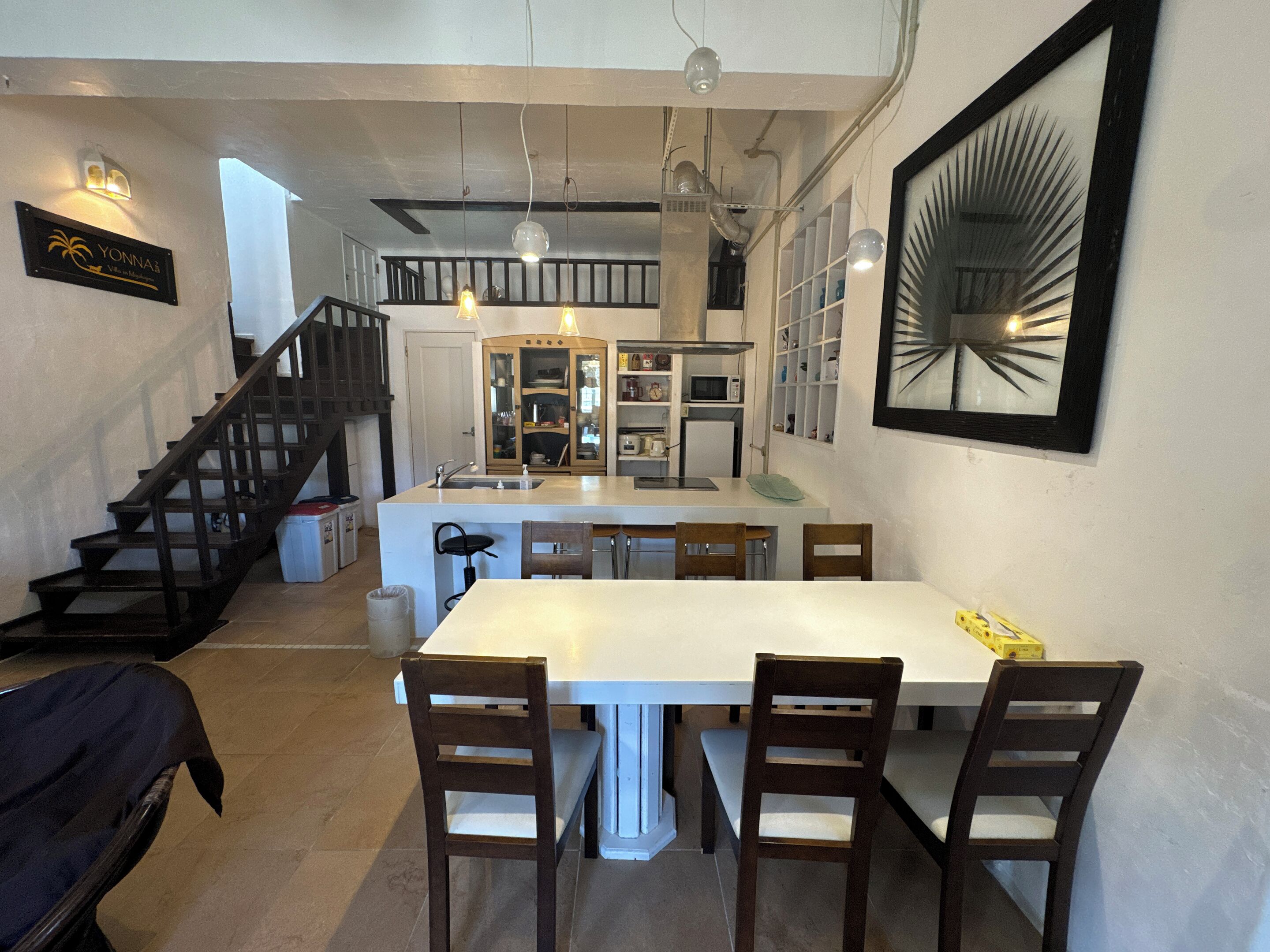 A modern kitchen with a central island, a dining area with four chairs, a staircase, and a framed artwork on the wall.