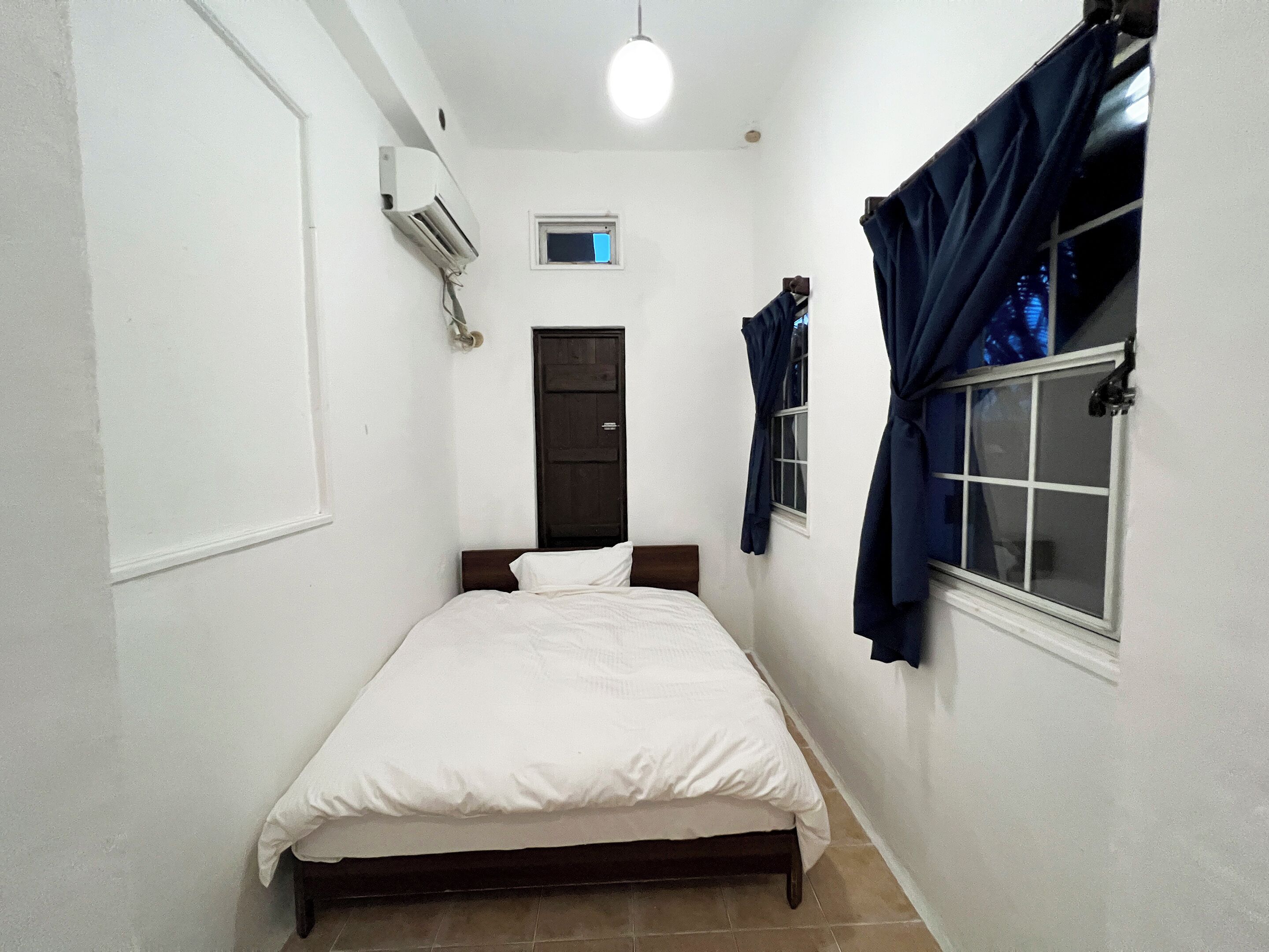 A narrow room with a bed, a window with curtains, a door, and an air conditioning unit.