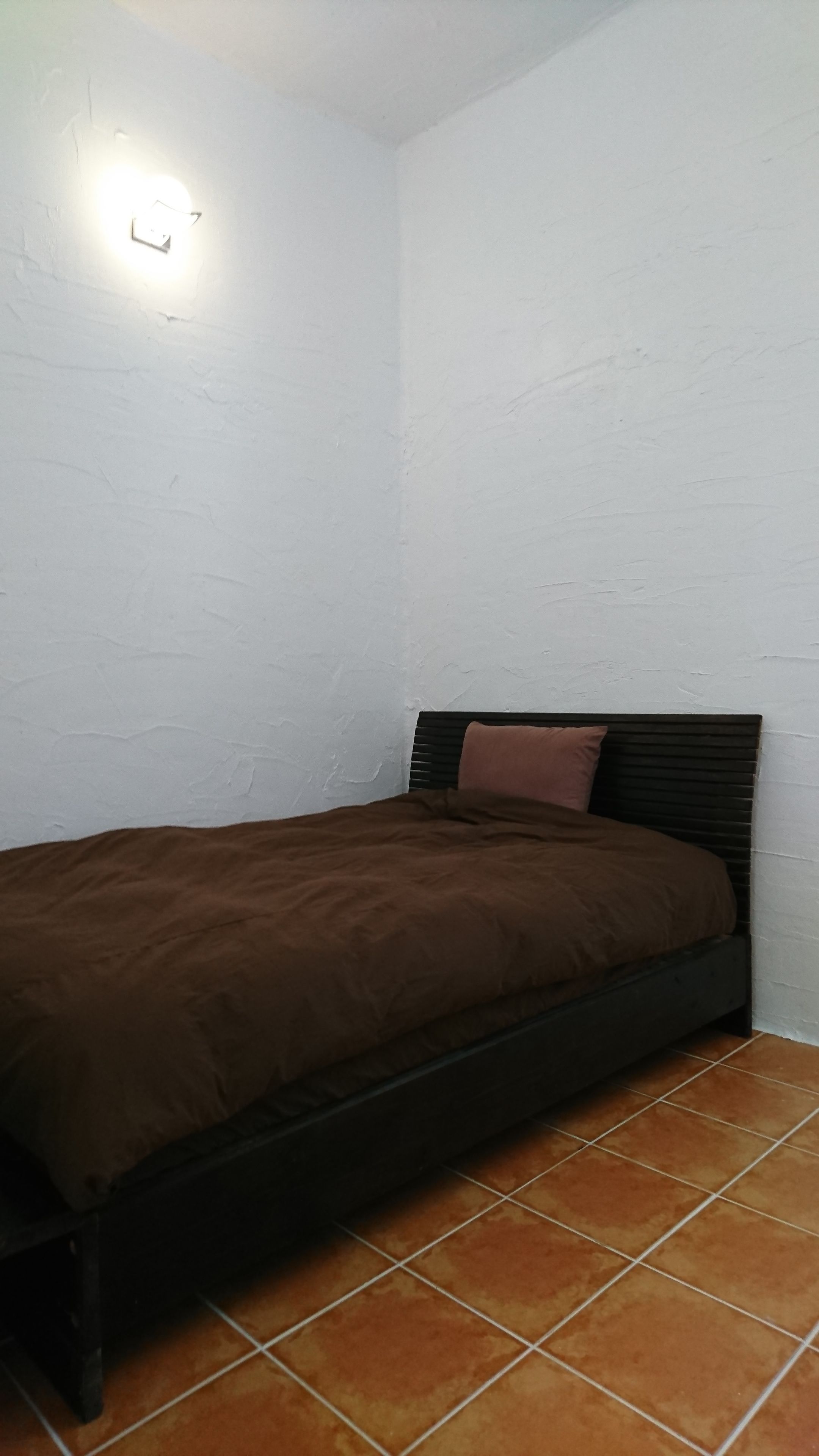 A single bed with a brown mattress and headboard, a white wall, and a tiled floor.