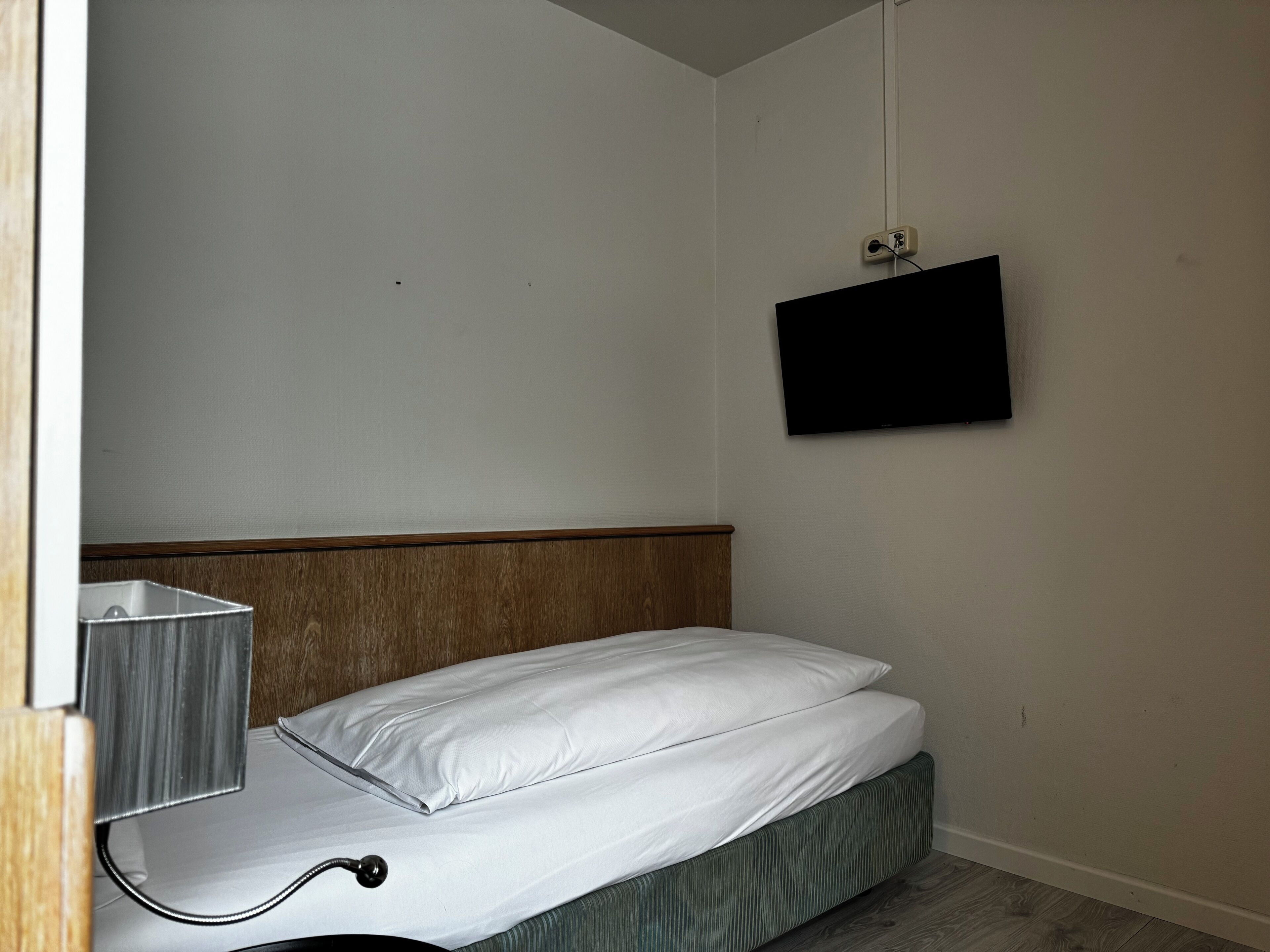Una habitación pequeña y sencilla con una cama, un televisor montado en la pared y un enchufe.