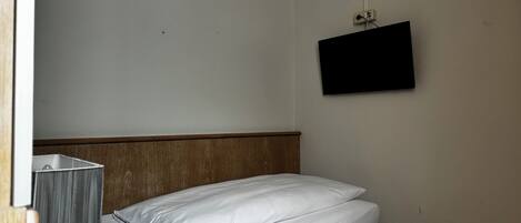 Una habitación pequeña y sencilla con una cama, un televisor montado en la pared y un enchufe.