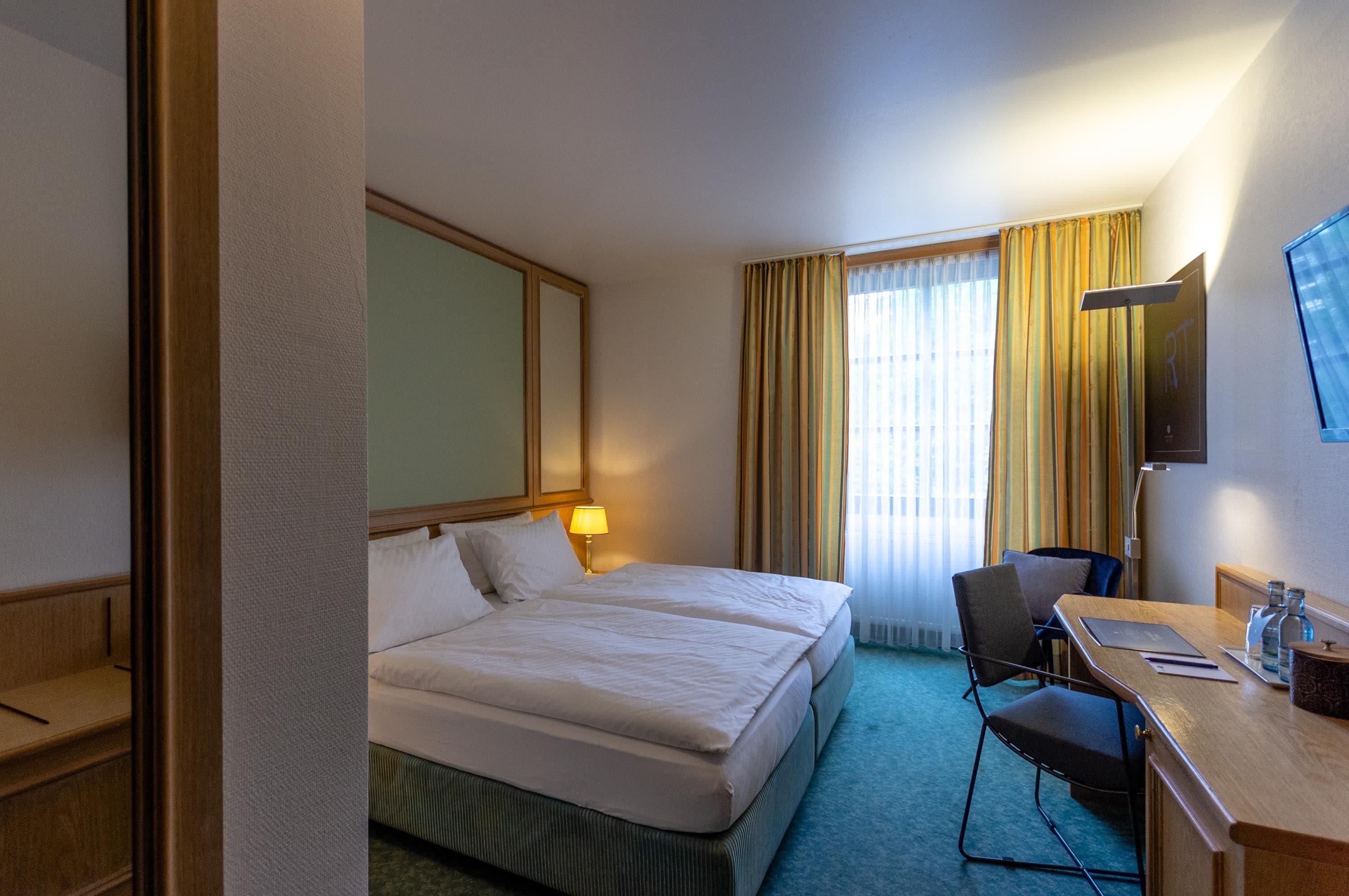 A hotel room with a double bed, a desk with a chair, a television, and a large window with curtains.