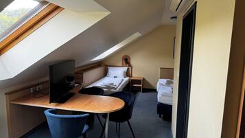 A compact hotel room with a sloped ceiling, a bed, a desk with a chair, a small bedside table, and a TV.
