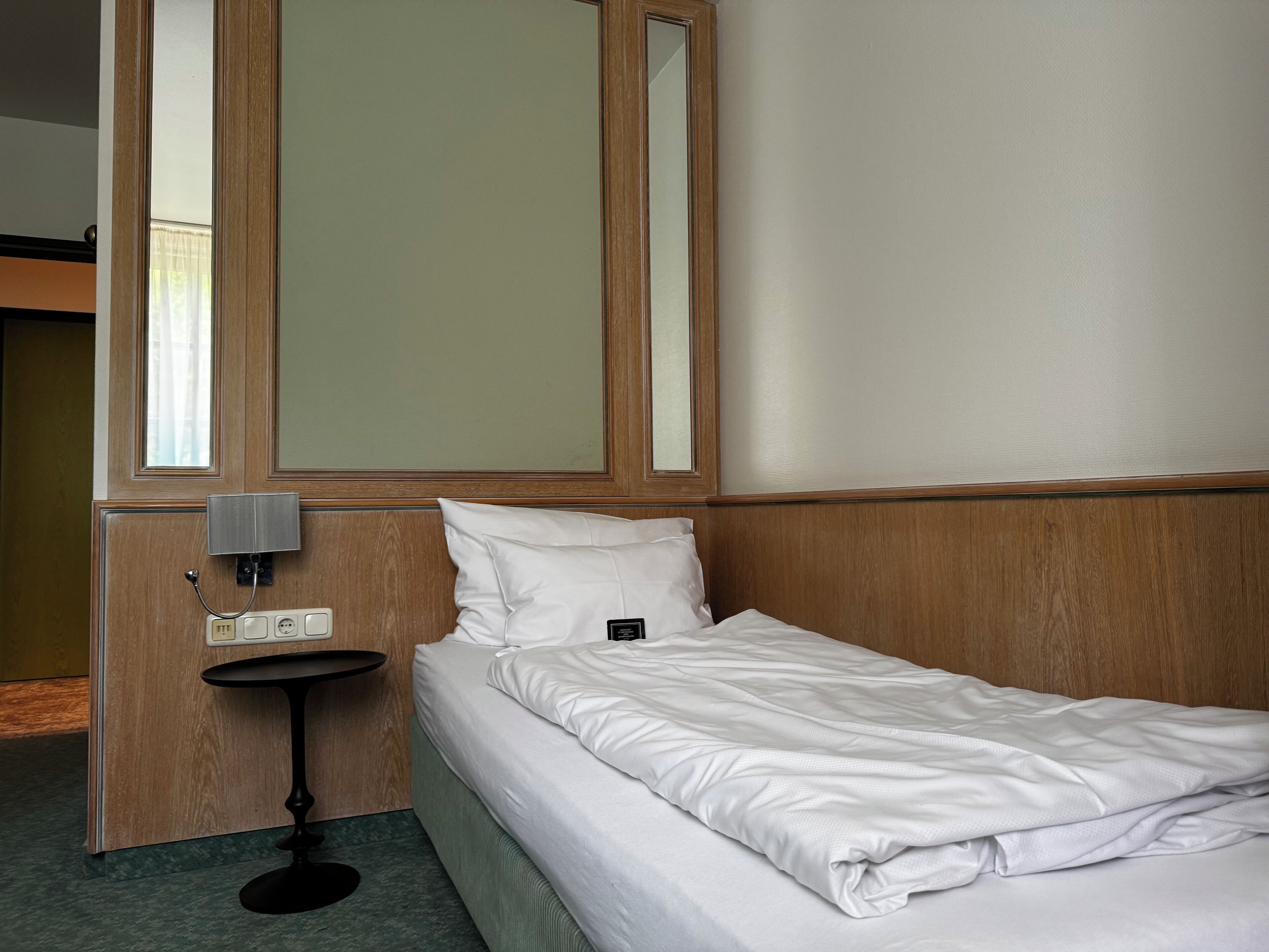 A hotel room with a single bed, a bedside table, a mirror, and a window with curtains.