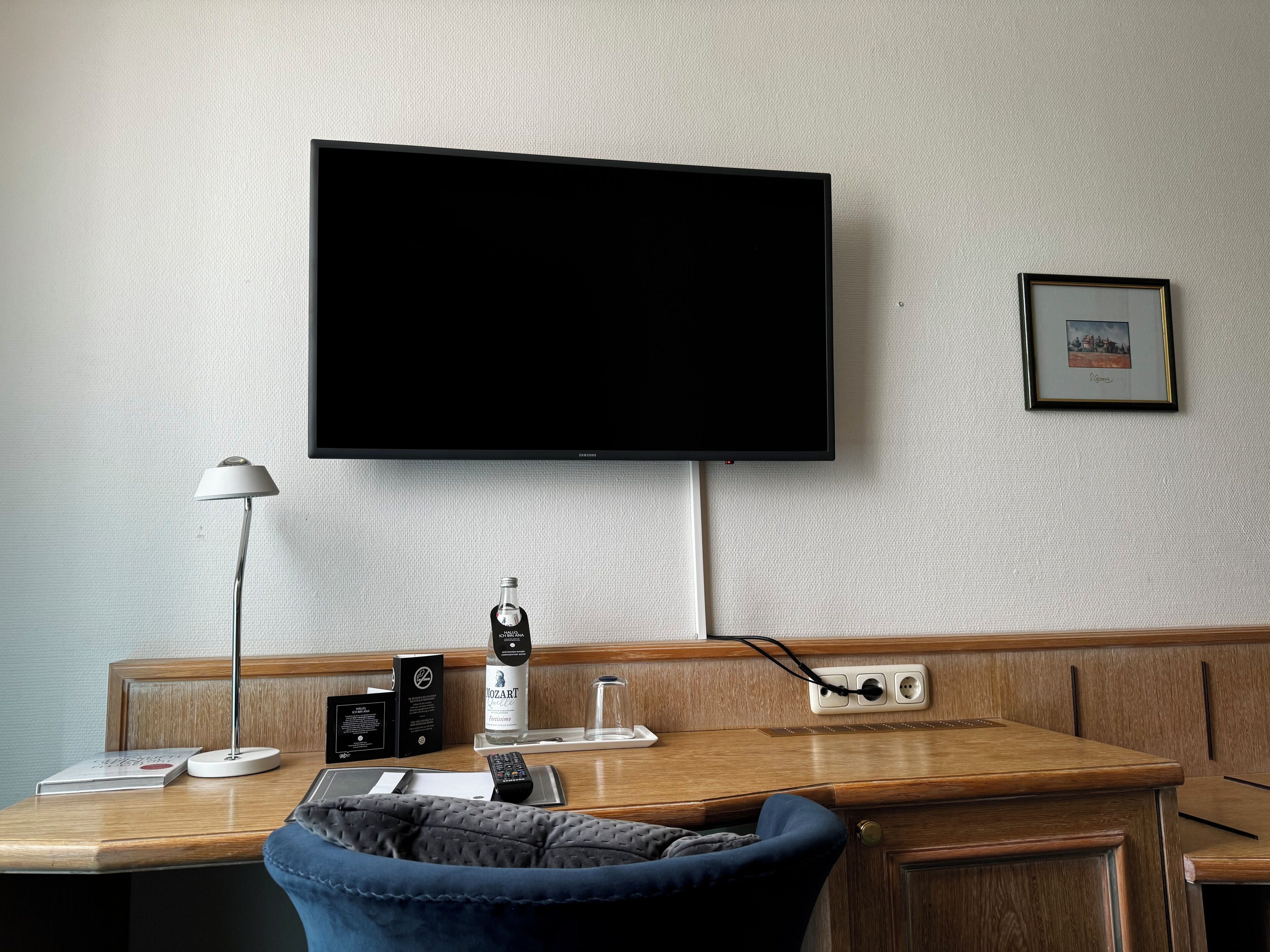 Une chambre d’hôtel avec un bureau en bois, un téléviseur à écran plat fixé au mur, un tableau encadré et une table de chevet avec une lampe, une bouteille et une télécommande.
