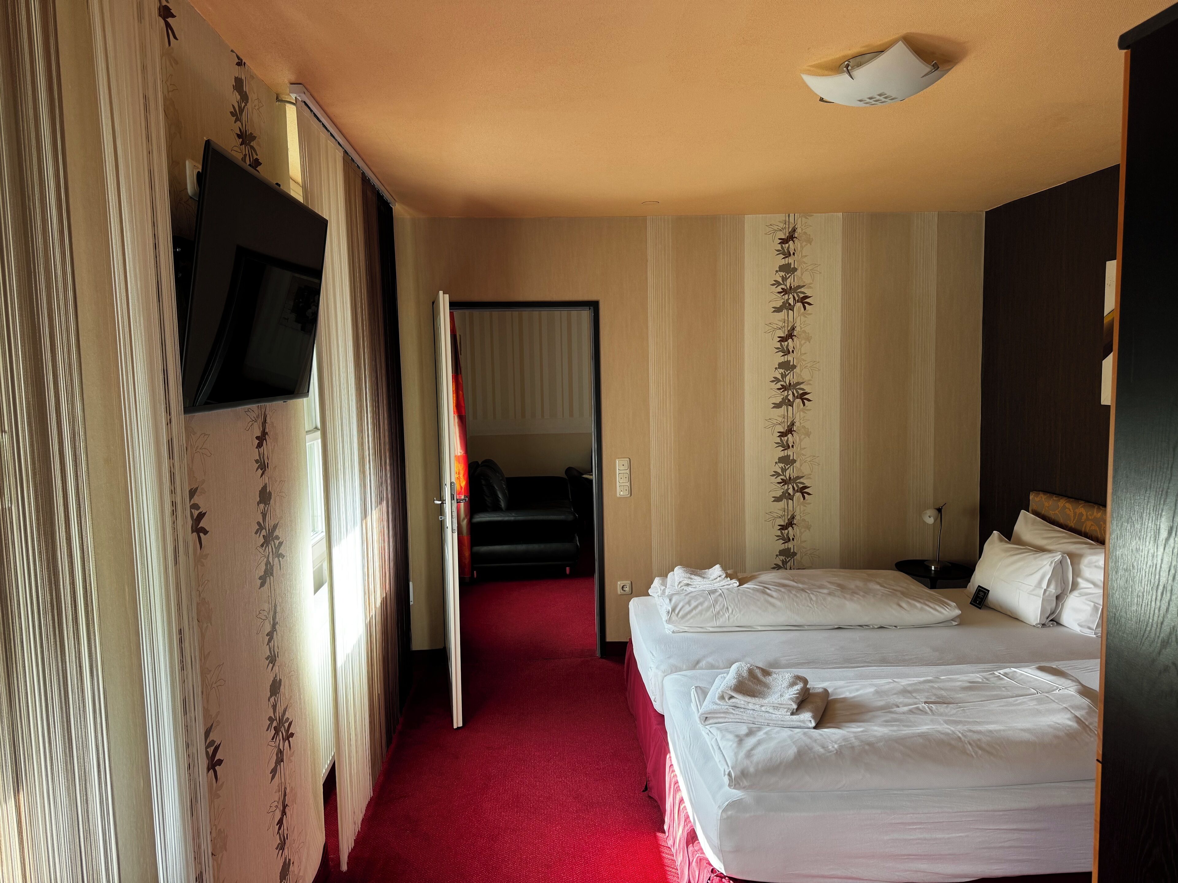 Habitación de hotel con dos camas, alfombra roja, televisor montado en la pared y una lámpara en el techo.