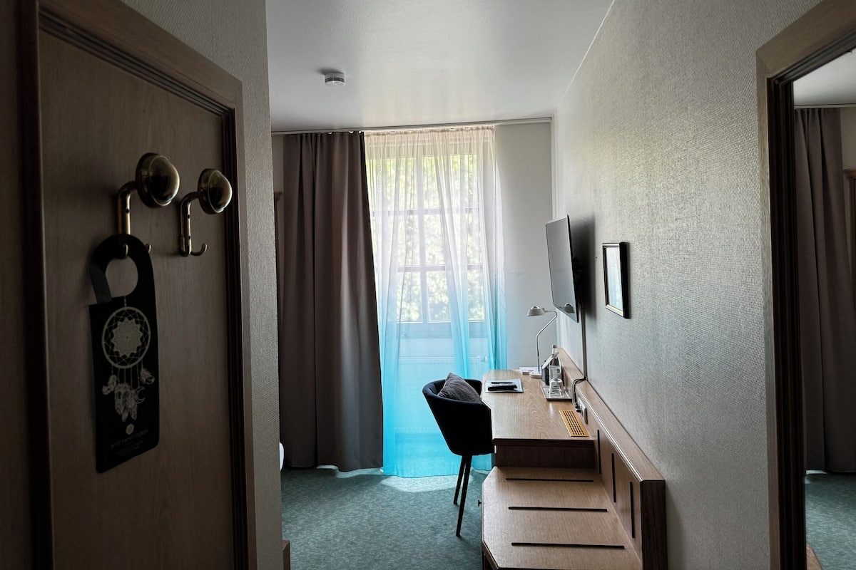 A hotel room with a wooden door, a desk with a chair, a TV mounted on the wall, and a window with curtains.